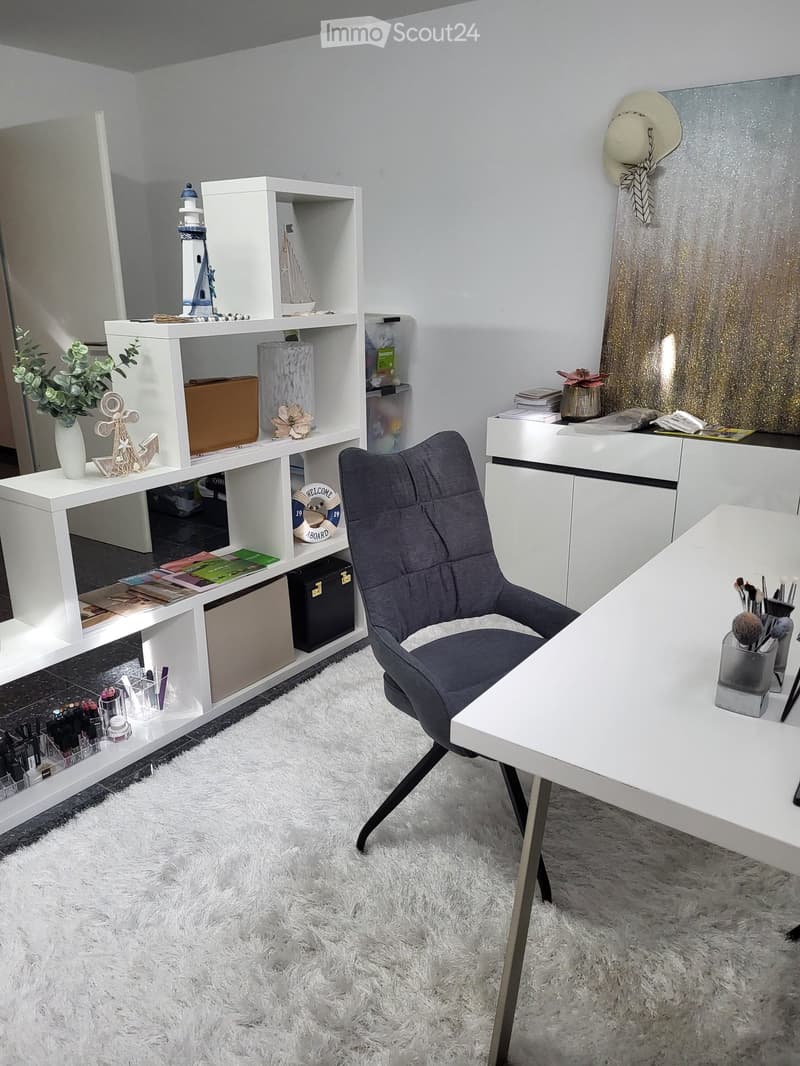 A well-organized room with a white shaggy rug, a grey chair, a white desk, and a white shelving unit holding books, a vase, and decorative items.