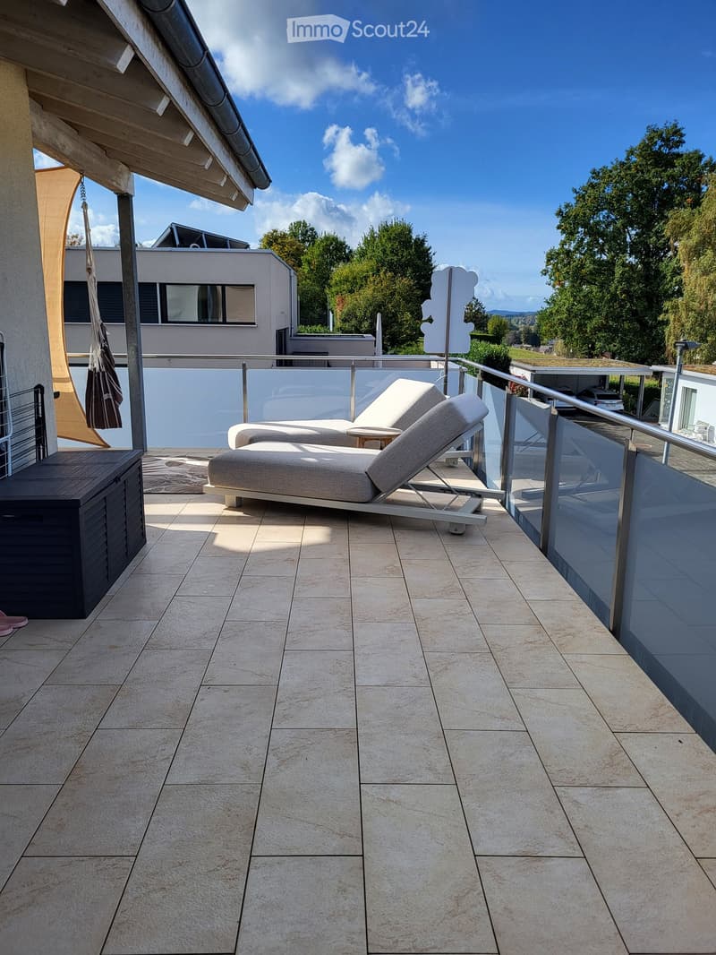 Modern balcony with beige tiles, sun loungers, small table, and storage box. Railings and outdoor umbrella visible.