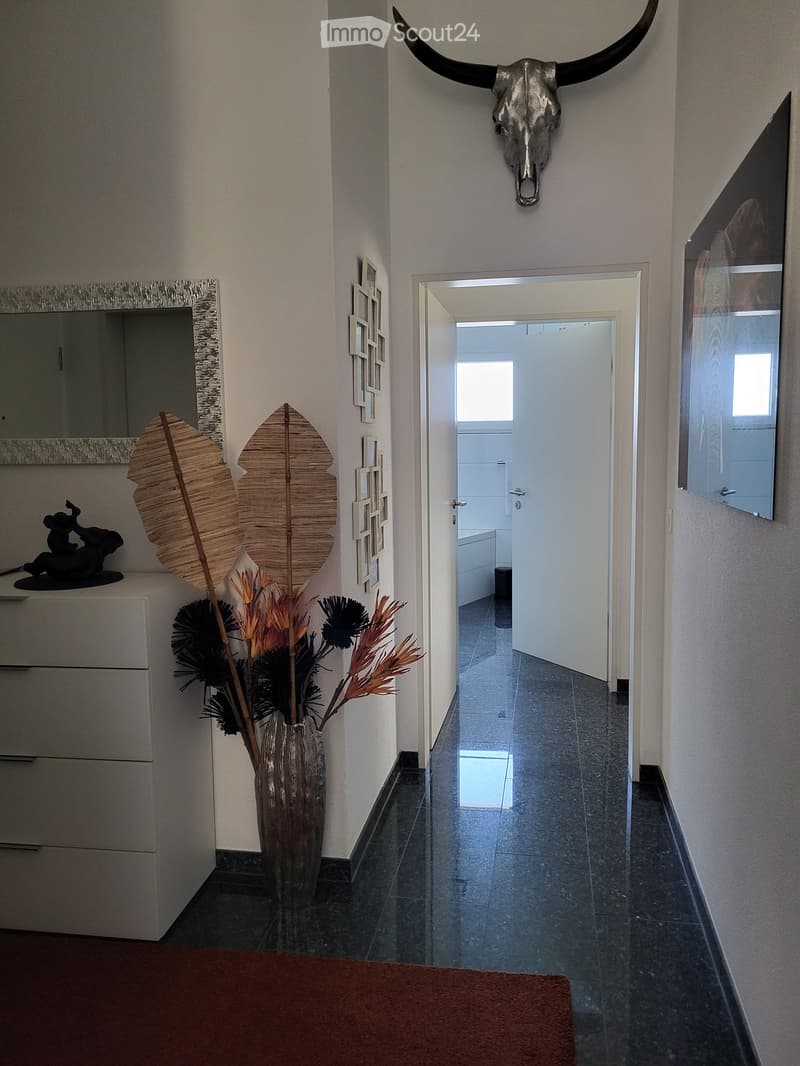 A modern hallway with dark tiled flooring, white walls, and decorative elements including a skull on the wall, a vase with dried flowers, and framed artwork. A doorway leads to another room.