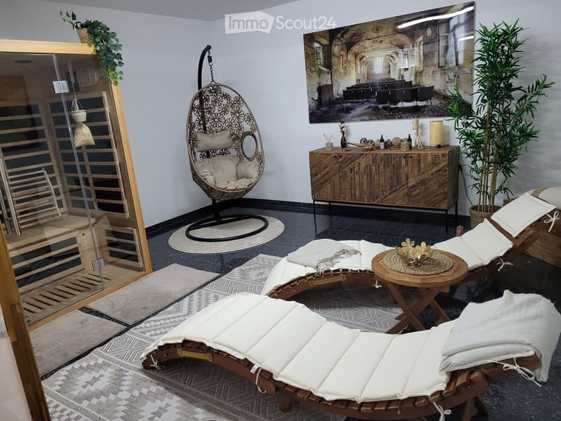 A modern, well-furnished room with a wicker hanging chair, wooden side table, wooden cabinet, and multiple wooden lounge chairs with white cushions. The room features a large mirror, a table runner, a table centerpiece, and potted plants, creating a cozy
