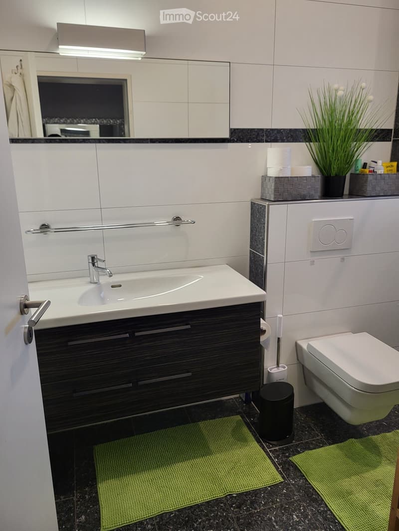 Modern bathroom with white tiles, dark wood cabinet, sink, mirror, toilet, and green rugs.