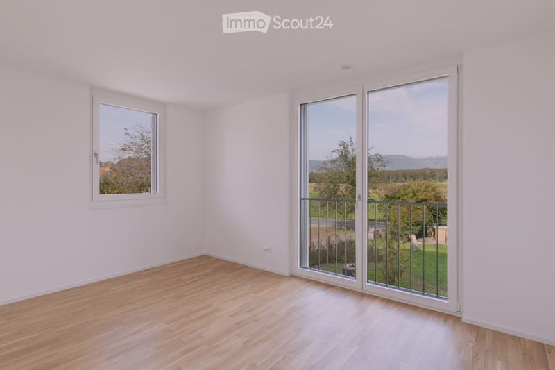 Empty room with wooden flooring, white walls, and large sliding glass doors leading to a balcony with a view of greenery and mountains.