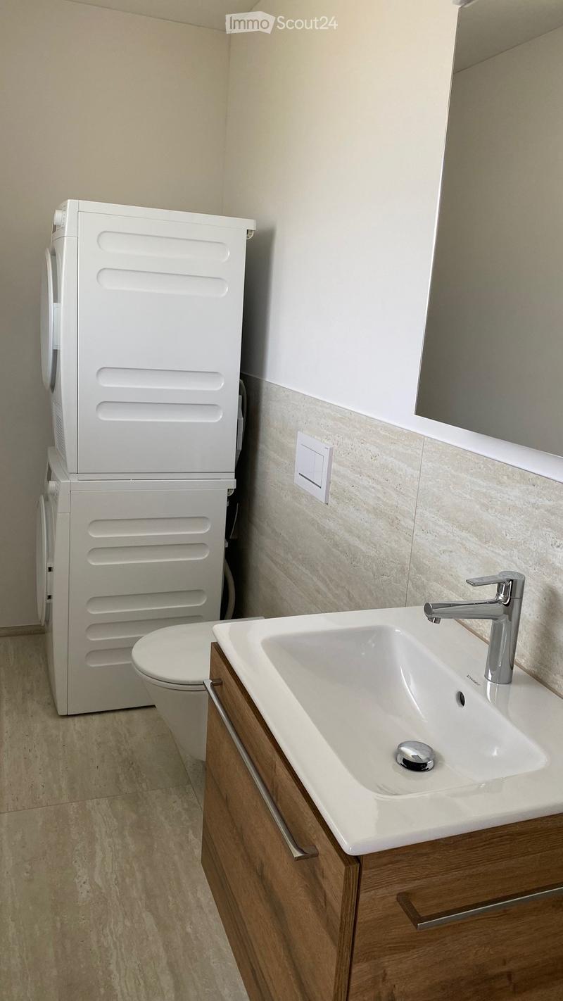 Modern bathroom with a white sink, chrome faucet, wooden cabinet, and toilet. Light-colored tiles and a mirror are visible.