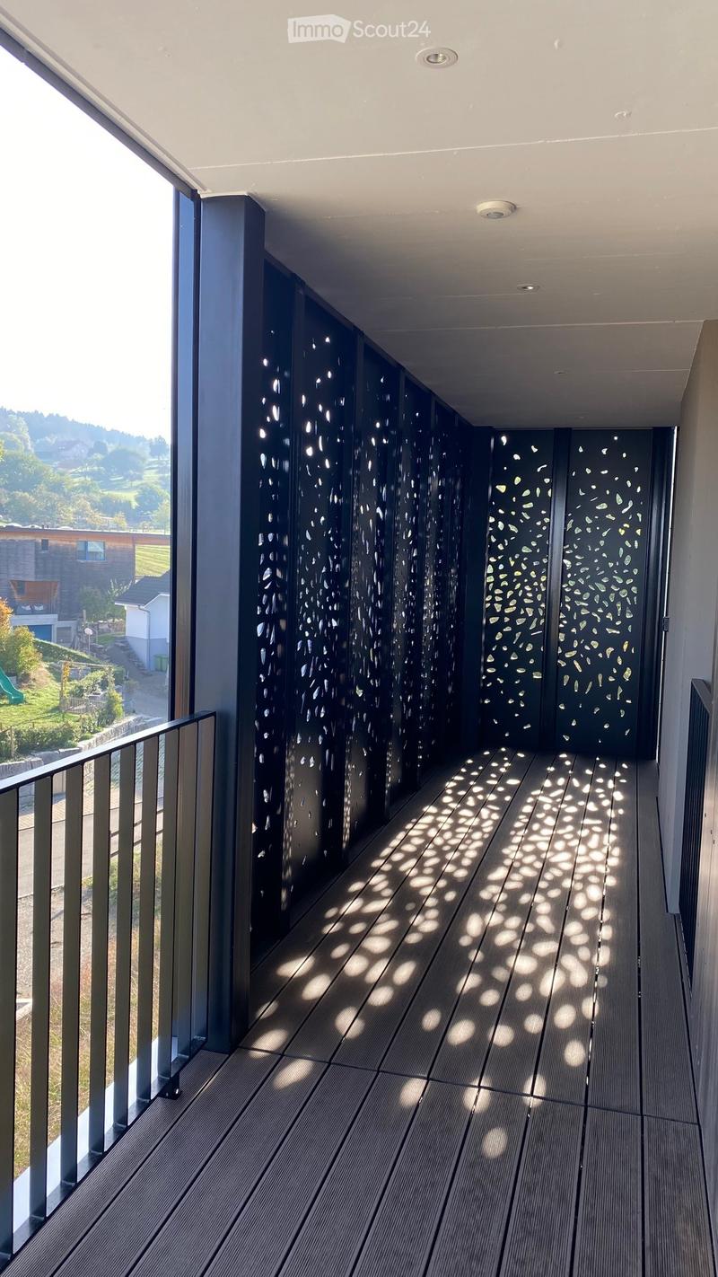 Modern balcony with wooden decking, dark privacy screens, and metal railing, overlooking a green landscape.