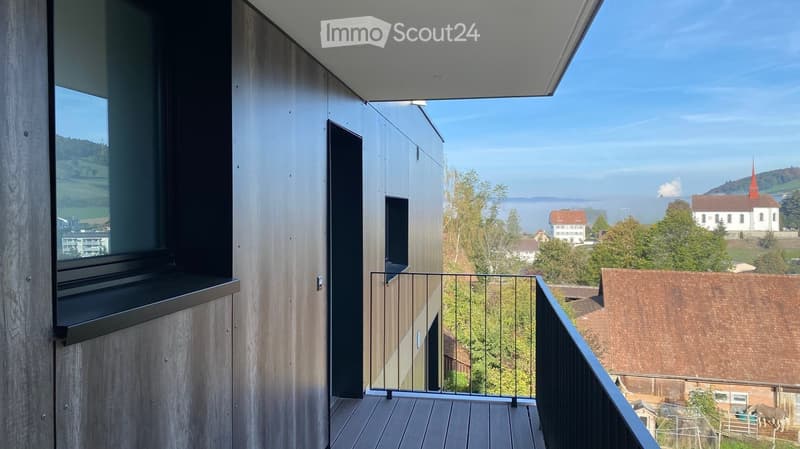 Modern balcony with wooden decking, glass railing, and expansive view of a town, church, and landscape.