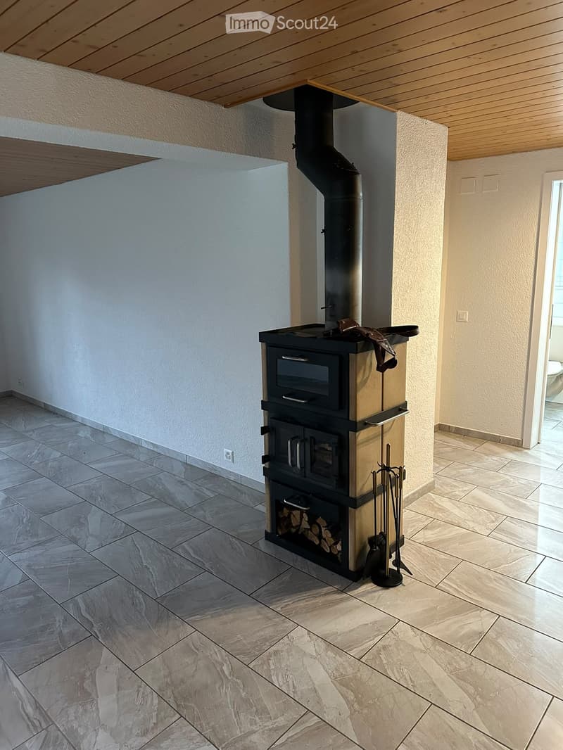 A modern, minimalist room with a large, black wood-burning stove as the focal point. The stove has a sleek design with a glass front and a storage compartment for firewood below. The floor is covered with large, light-colored tiles, and the walls are pain