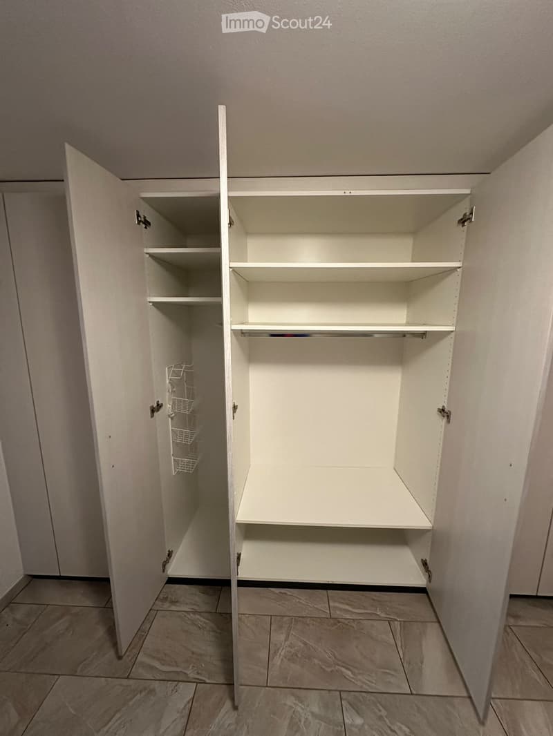 Empty closet with white shelves and a wire basket on the left side. The floor is tiled, and the walls are painted a light neutral color.
