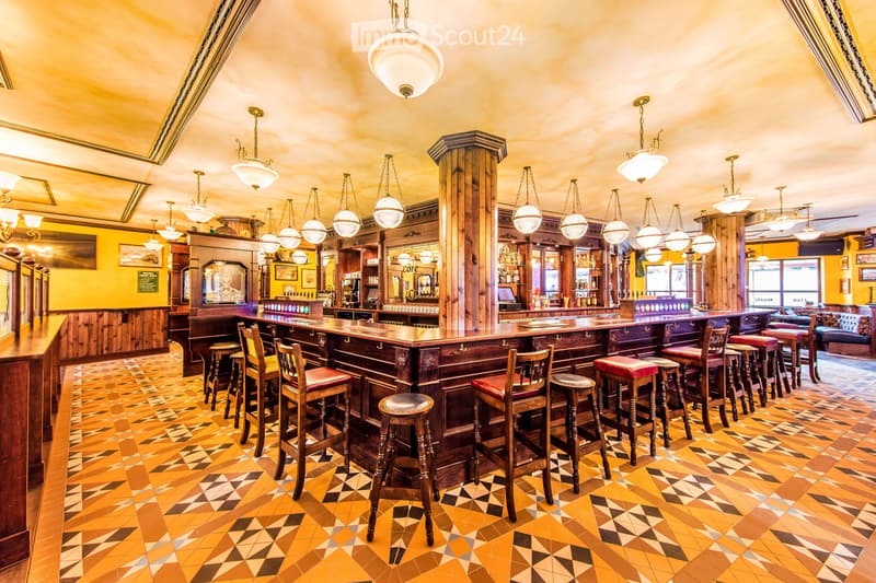 A well-furnished bar area with a long wooden bar counter, multiple bar stools, pendant lights, shelves stocked with bottles, and a cozy seating area with a sofa.