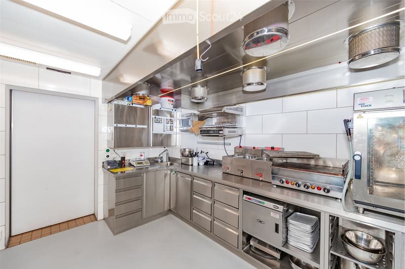 Commercial kitchen with stainless steel appliances, including a refrigerator, microwave, oven, stove, fryers, sink, and dishwasher. The space features stainless steel cabinets, drawers, and a countertop with various kitchen tools and equipment.