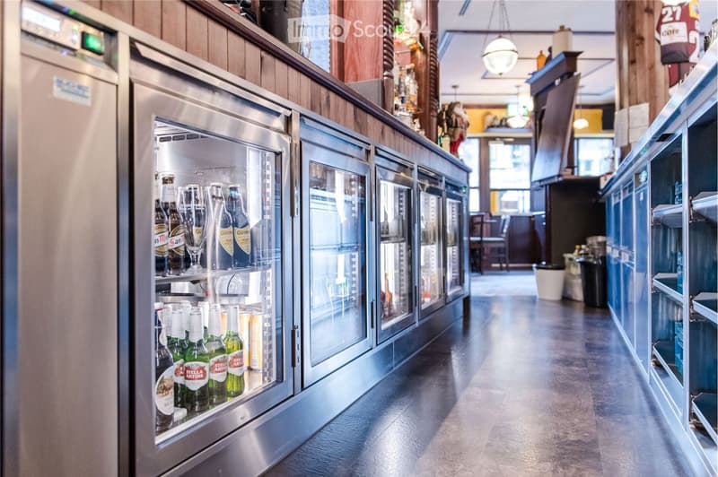 A modern bar area with stainless steel refrigerators stocked with various beer bottles and glasses, wooden counter, and hanging light fixture.