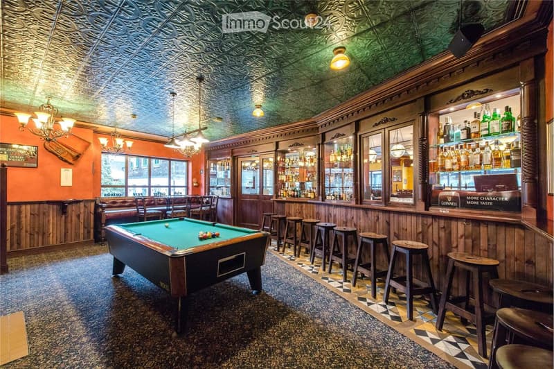 A stylish bar area with a pool table, wooden bar, shelves of bottles, and several bar stools. The ceiling features ornate tinwork and chandeliers, and the walls are painted in warm orange tones with wooden paneling.