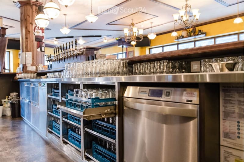 A modern bar area with a stainless steel counter, multiple beer taps, neatly stacked glasses, a dishwasher, and storage cabinets. The space is well-lit with hanging chandeliers and has a clean, organized appearance.
