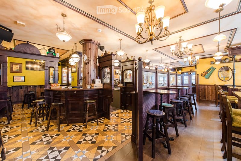 A cozy bar area with wooden bar counters, multiple stools, tables, chairs, and ornate chandeliers. The decor includes framed pictures and a mirror on the wall.