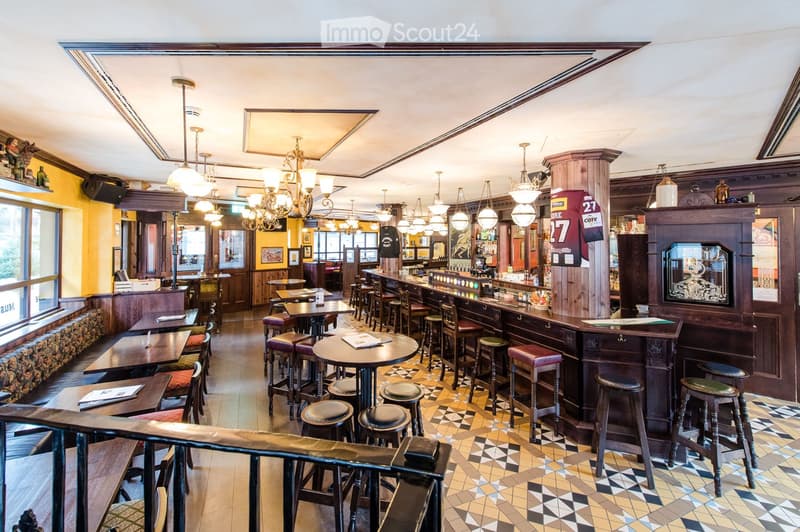 A well-lit pub interior with wooden tables, bar counter, and multiple stools. The space features ornate chandeliers, a patterned tile floor, and a mix of seating options including tables and bar stools.