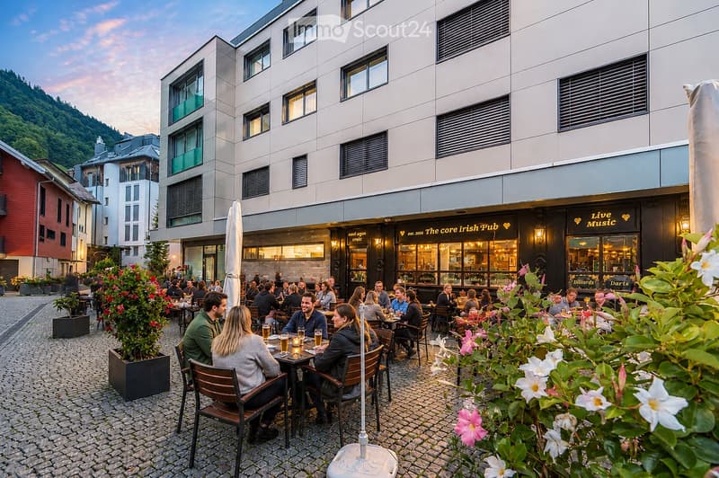 An outdoor seating area of a pub with people enjoying drinks and conversation, surrounded by tables, chairs, and potted plants, with a modern building in the background.