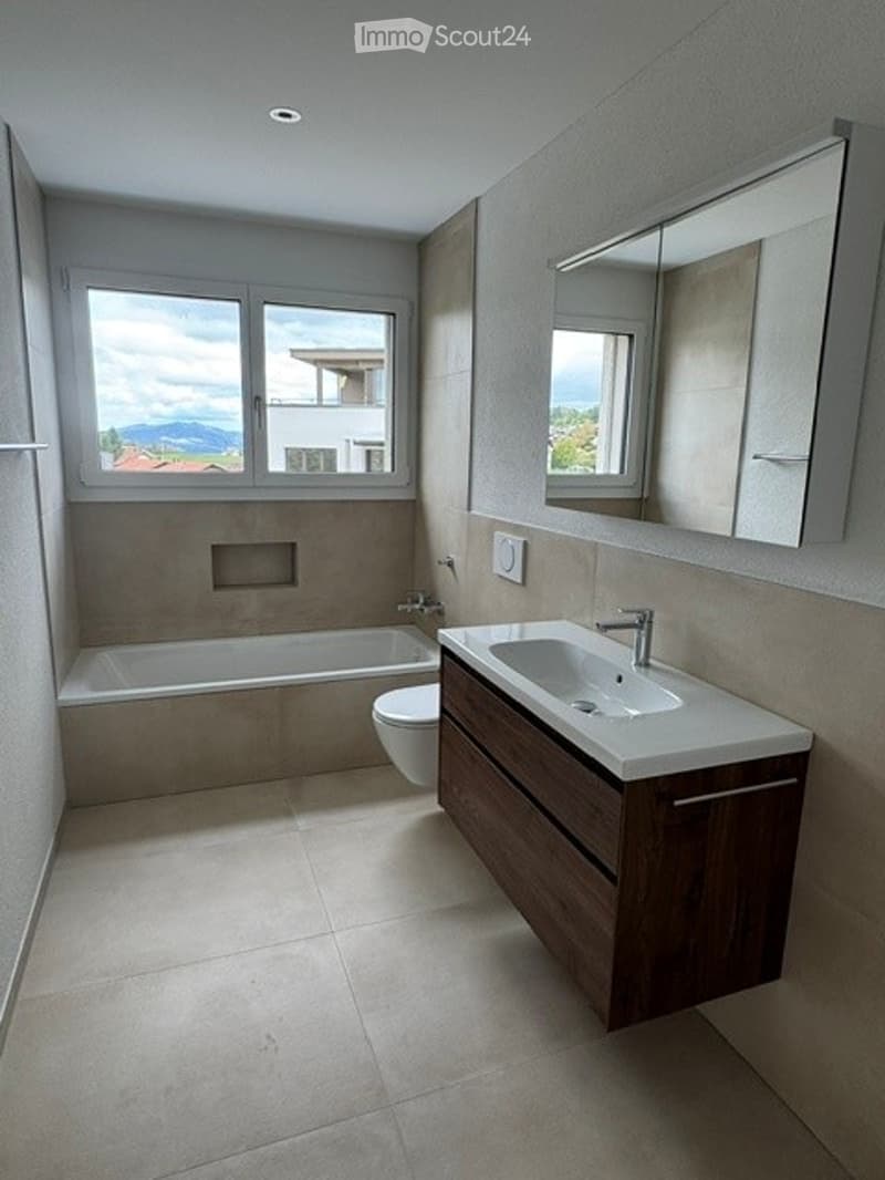 Modern bathroom with a bathtub, toilet, sink, and large mirror. Natural light from windows offers a scenic view.