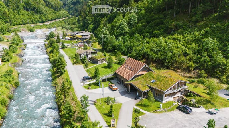 Aerial view of a rural building complex with a garage, parking area, and surrounding greenery near a river.