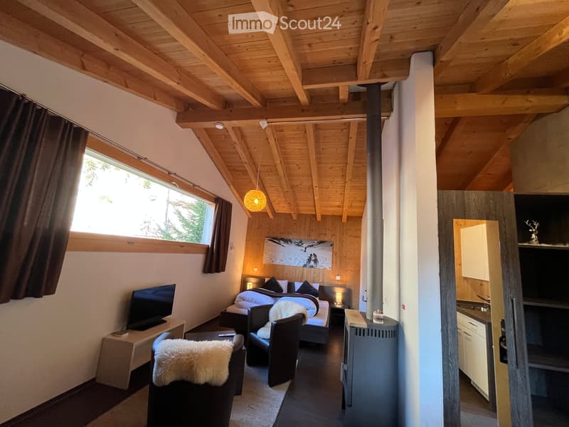 Cozy, well-furnished room with exposed wooden beams, a double bed, two chairs, a television, a small table, and a lamp. The room features large windows with curtains and a view of greenery.