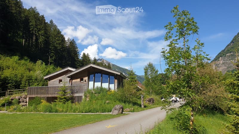 A modern wooden house with large windows and a balcony, surrounded by lush greenery and mountains in the background.