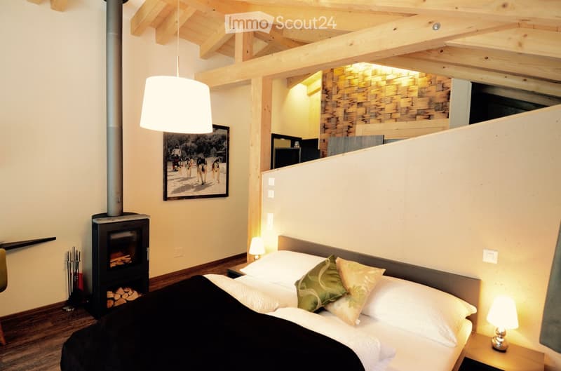 Cozy bedroom with wooden beams, a fireplace, a large bed with pillows and a blanket, nightstands with lamps, and a picture frame on the wall.