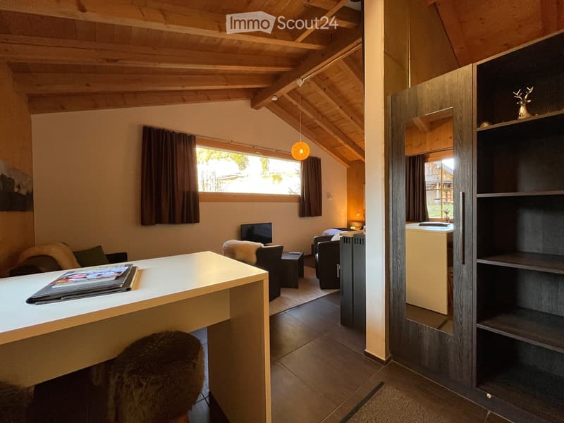 A cozy, well-lit room with wooden beams, a large window, a desk, a chair, a small table, and a shelf with decorative items.