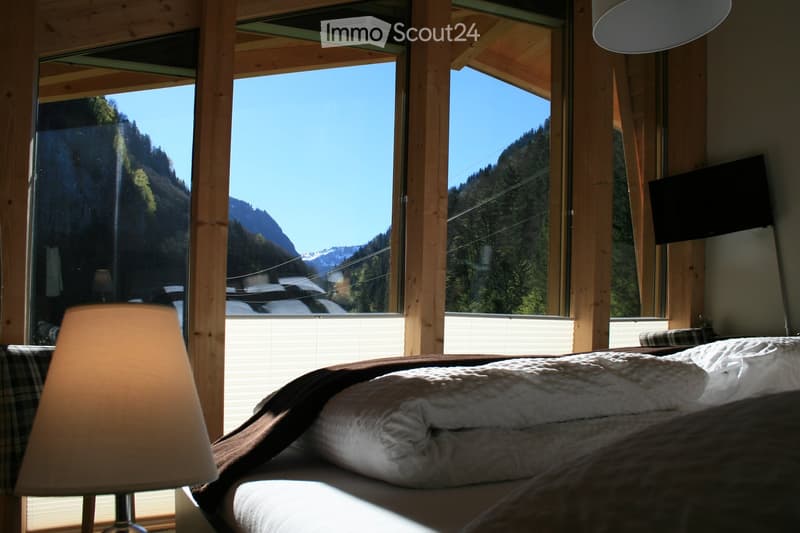 Bright bedroom with large windows offering a scenic mountain view, featuring a neatly made bed with pillows and blankets, a bedside lamp, and a mounted television.