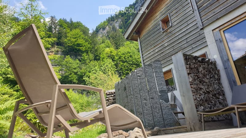 Wooden house with a stone chimney, surrounded by greenery and a stack of firewood. A single lounge chair is placed on a wooden deck.