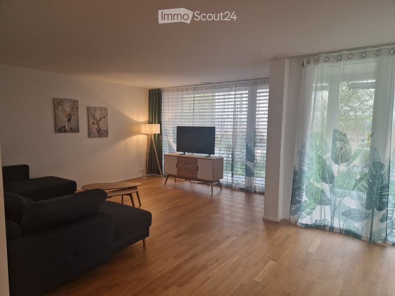 Modern living room with wooden flooring, large windows with blinds, a dark grey sofa, a wooden coffee table, a TV on a white cabinet, and a floor lamp.