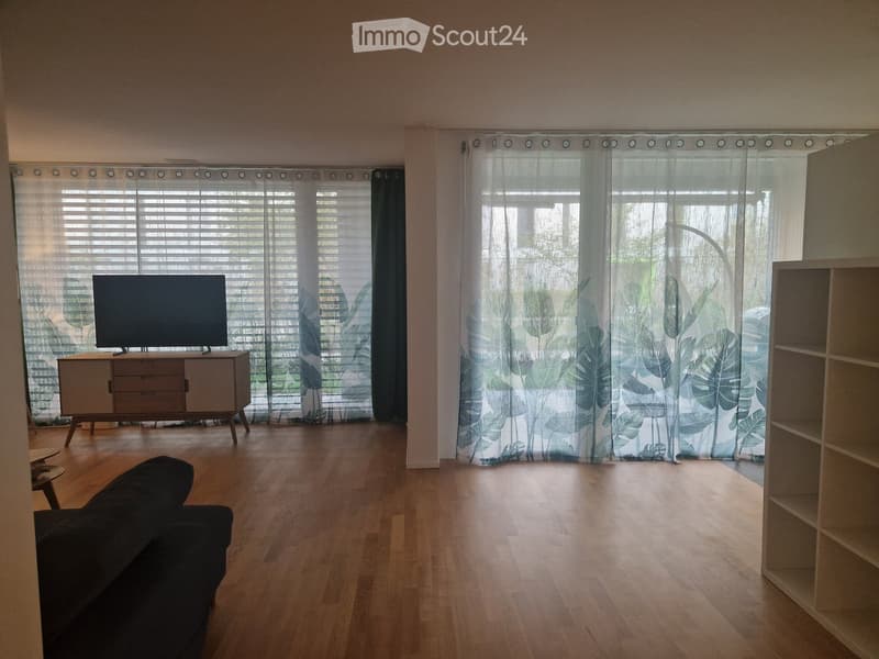 Living room with wooden flooring, large windows with blinds and curtains, a black sofa, a television on a cabinet, and a shelf unit.