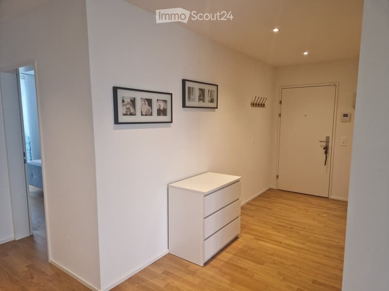A clean, unfurnished hallway with light wooden flooring, white walls, and a small white cabinet. Two framed pictures are mounted on the wall, and a coat rack is visible near the door.