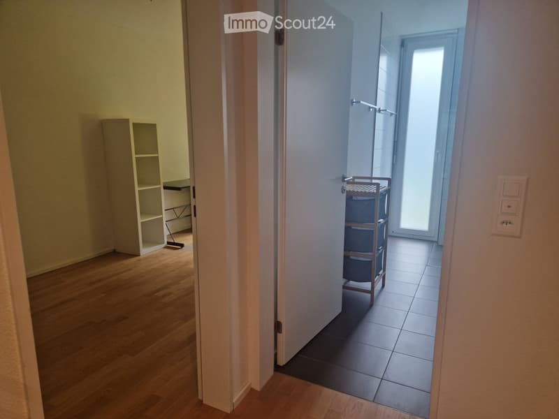 Empty hallway with wooden flooring, white walls, and a small shelf on the left side. A door leads to a tiled room with a small cart inside.