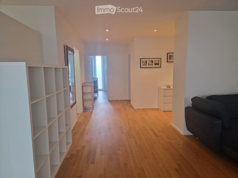 A well-lit hallway with wooden flooring, a black sofa on the right, a white cabinet, a mirror, and a small pink chair visible in the background.