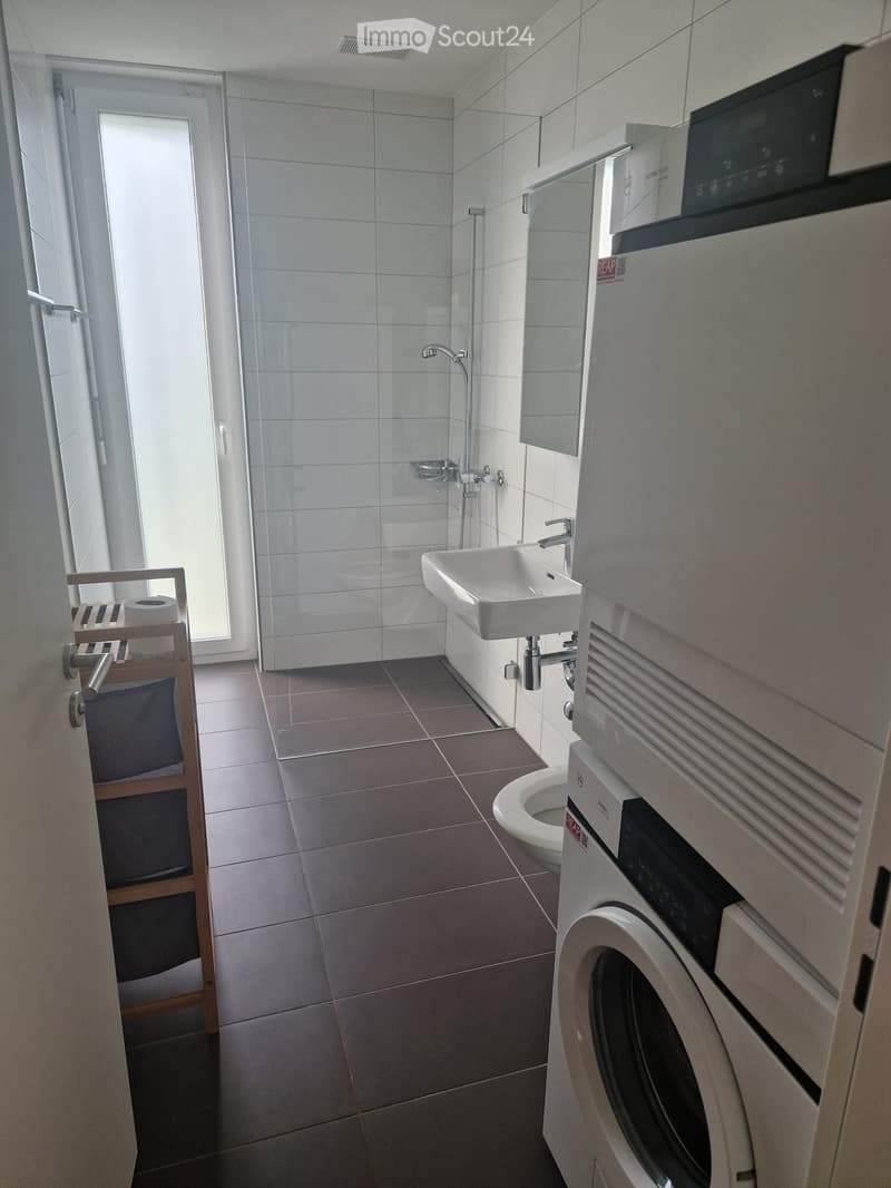 Modern bathroom with a white sink, toilet, glass-enclosed shower, and a washing machine. The space features tiled walls and floor, a towel rack, and a window providing natural light.
