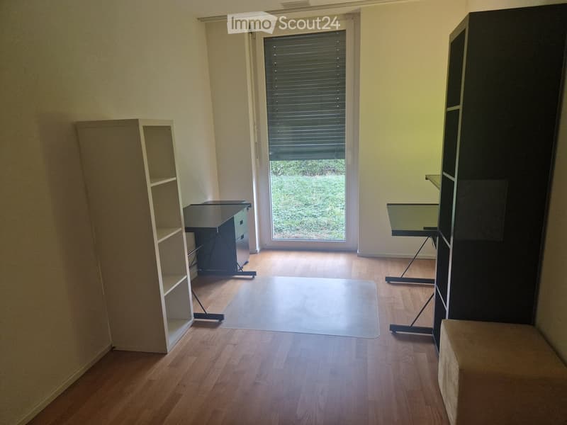 A small, unfurnished room with wooden flooring, a desk, a shelf, and a chair. The room has a large window with blinds, allowing natural light to enter.