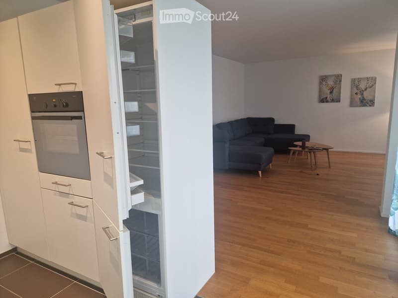 Modern living room and kitchen area with a dark blue sofa, coffee table, and white kitchen cabinets with stainless steel appliances.