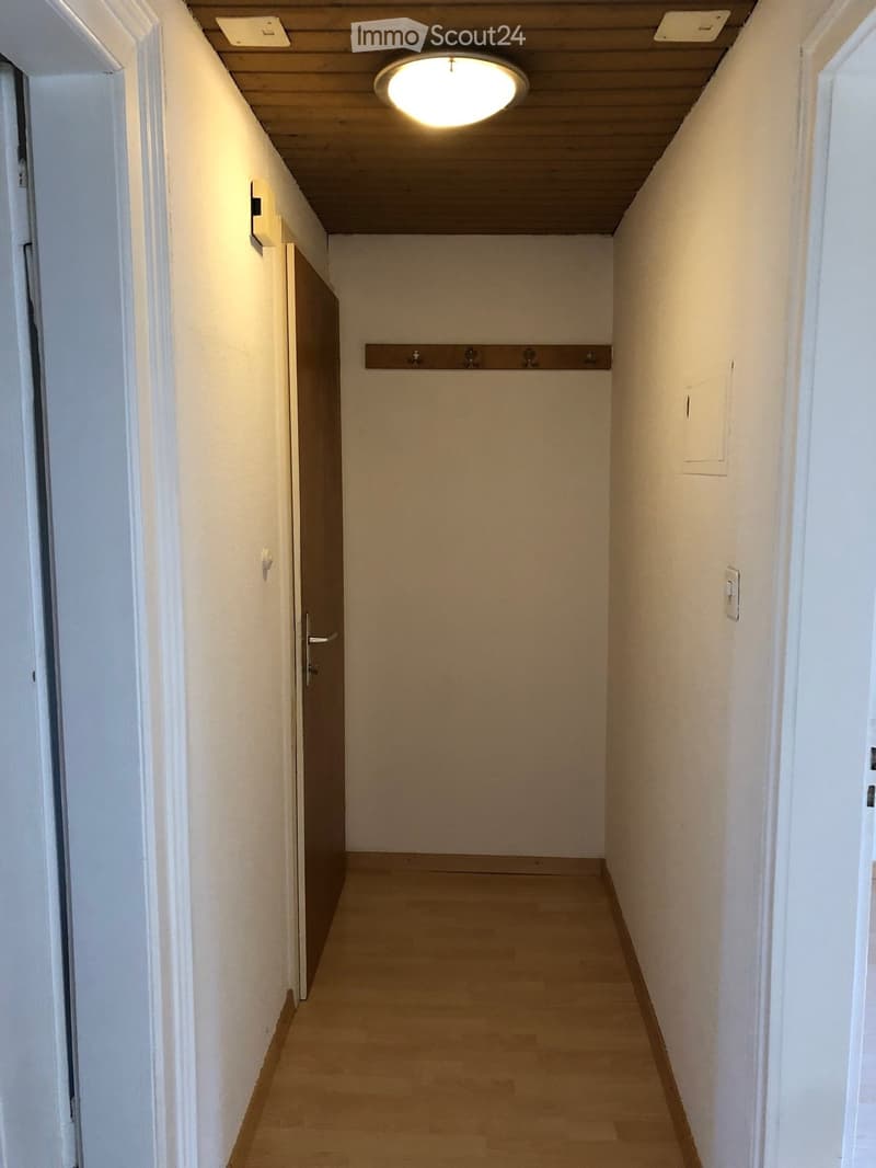 Empty hallway with wooden flooring, white walls, and a closed door on the left side. The ceiling has recessed lighting and a wooden panel design.