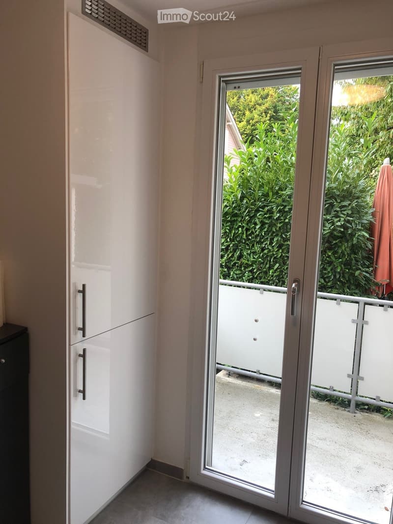 A narrow hallway with a sliding glass door leading to a balcony or terrace, featuring a white cabinet on the left and a view of greenery outside.