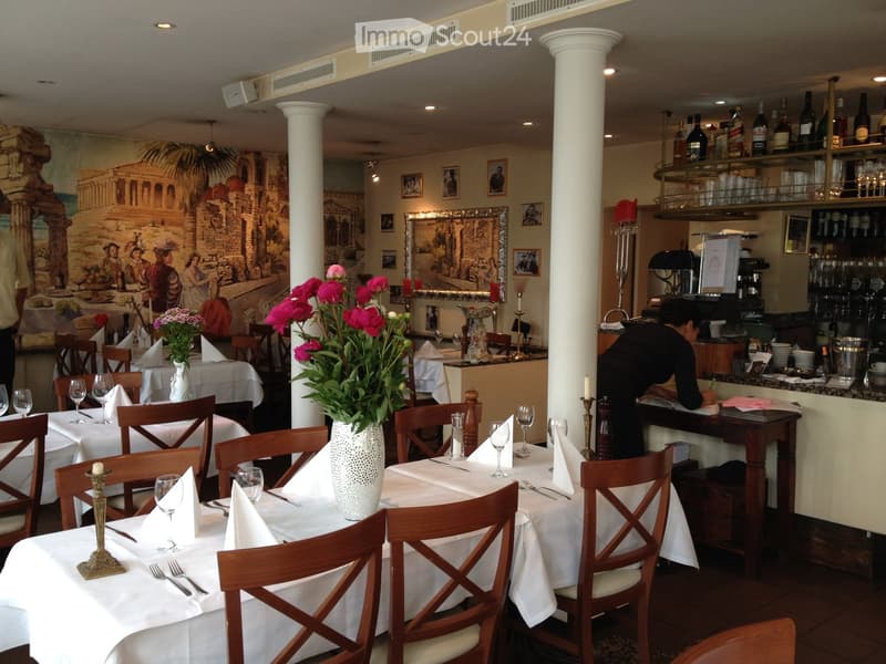 Elegant restaurant interior with white tablecloths, wooden chairs, decorative vases, wine glasses, and shelves of bottles and glasses.