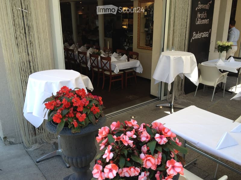 Outdoor seating area with tables, chairs, and flower pots, adjacent to a restaurant interior.