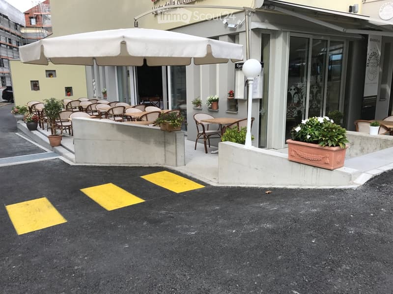 Outdoor seating area of a restaurant with tables, chairs, potted plants, and a large umbrella. The area is raised slightly above street level with concrete barriers and decorative planters.