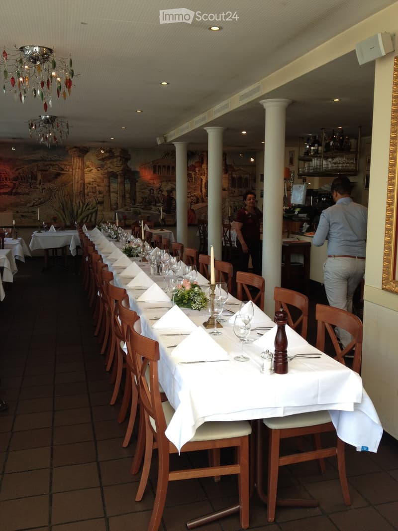 Restaurant dining area with long tables set for guests, featuring white tablecloths, napkins, wine glasses, and candles. Wooden chairs surround the tables, and a bar area is visible in the background.