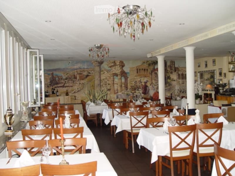 Restaurant interior with multiple tables and chairs, each set with white tablecloths, candles, and glassware. Large chandeliers hang from the ceiling, and the walls feature a painted mural depicting classical scenes. The room has large windows on one side