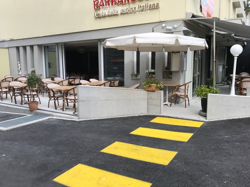 Outdoor seating area of a restaurant with tables, chairs, and an umbrella, surrounded by potted plants and a building entrance.