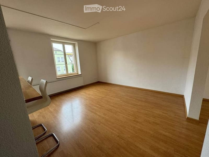 Empty room with wooden flooring, white walls, and a single window. A small table and chairs are positioned near the window.