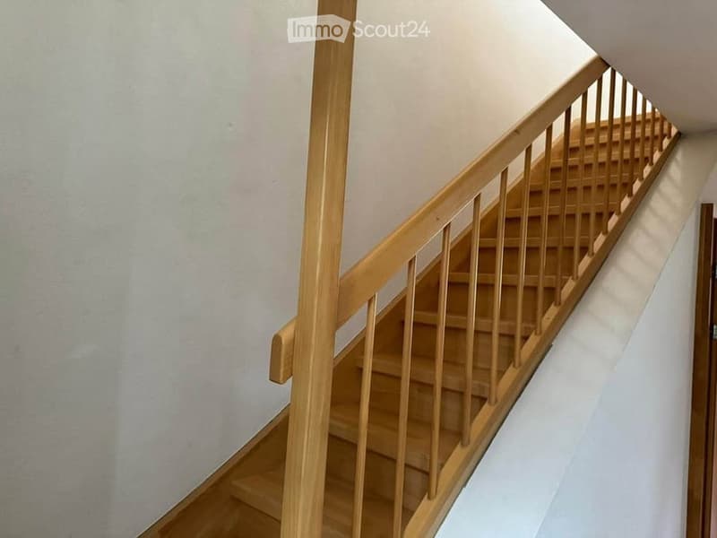 Wooden staircase with a handrail and open balustrade, leading upwards in a well-lit interior space.