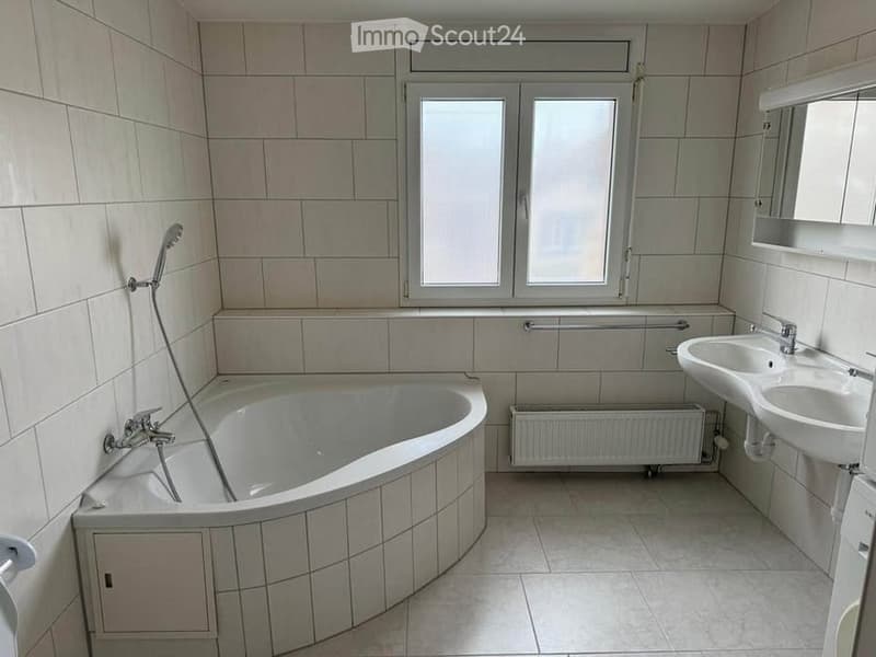Modern bathroom with a corner bathtub, double sink, tiled walls and floor, and a window above the sink.