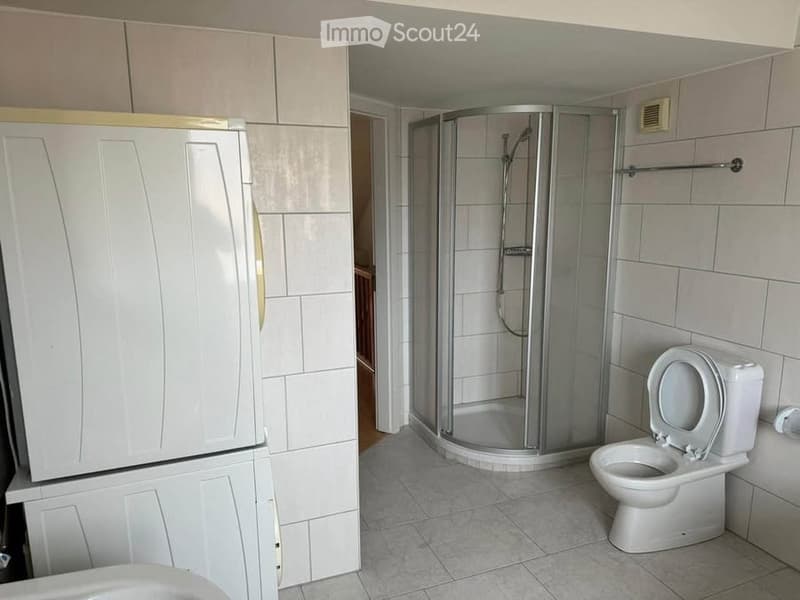 Modern bathroom with white tiled walls and floor, featuring a toilet, a glass-enclosed shower stall, and a white cabinet with gold handles.