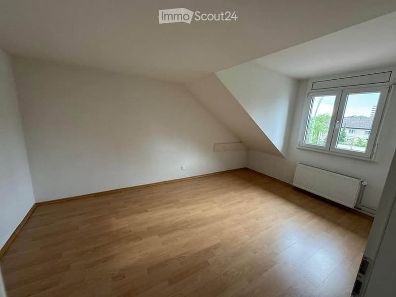 Empty attic room with wooden flooring, white walls, and a large window offering natural light. The room features a slanted ceiling and a radiator under the window.
