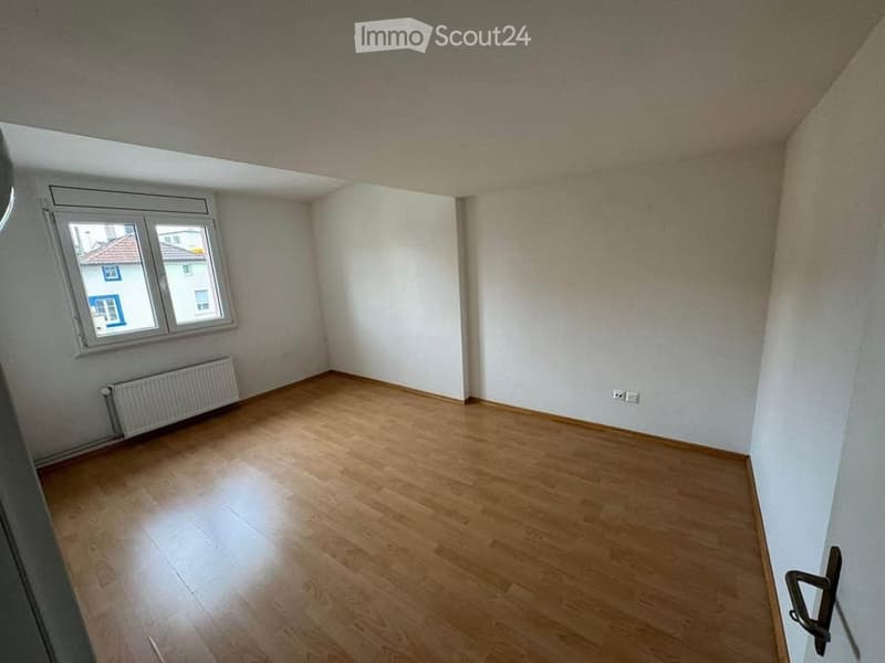 Empty room with wooden flooring, white walls, and a window with a view of a neighboring building.
