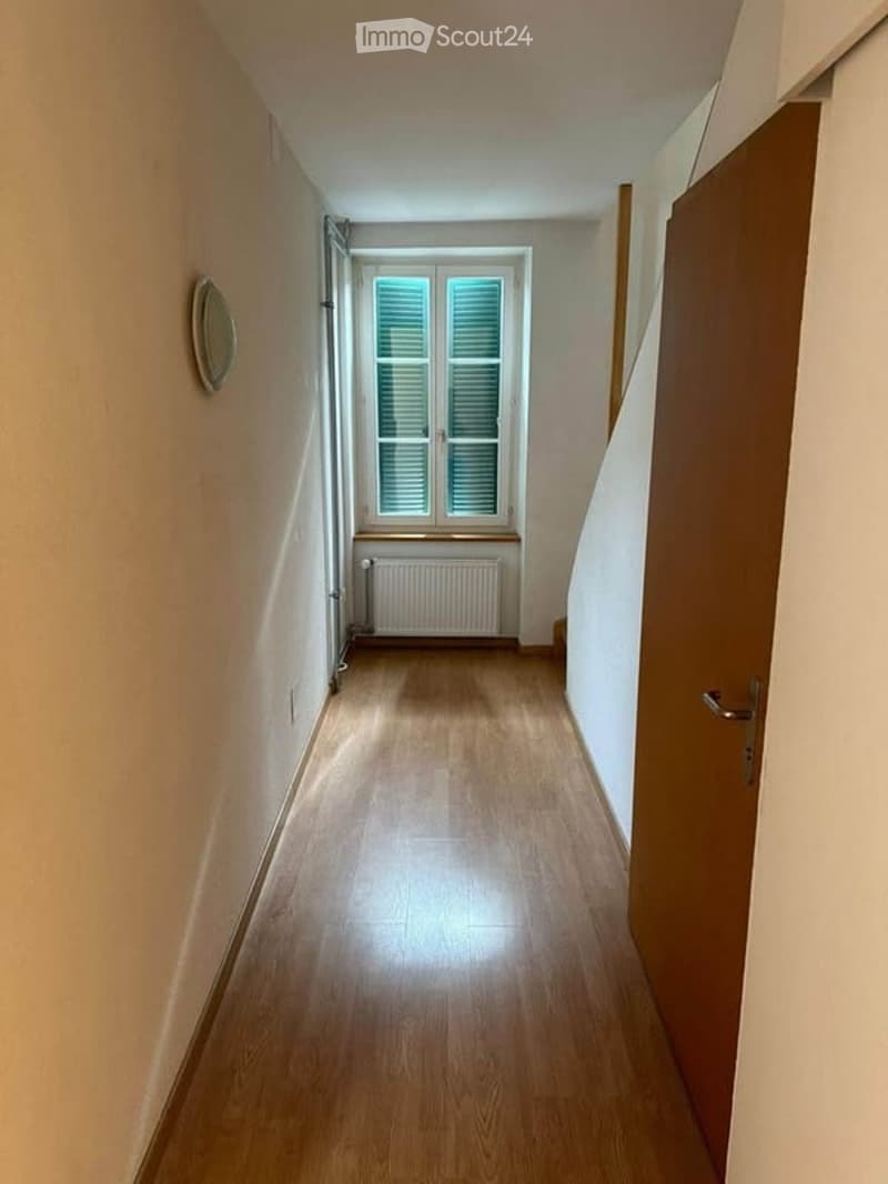 A narrow hallway with wooden flooring, white walls, and a window at the end. A radiator is placed below the window, and a closed wooden door is visible on the right side.